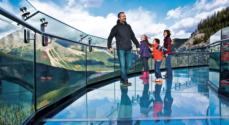 A family standing on the glass floor of the Columbia Icefield Skywalk with mountains reflected in the panels